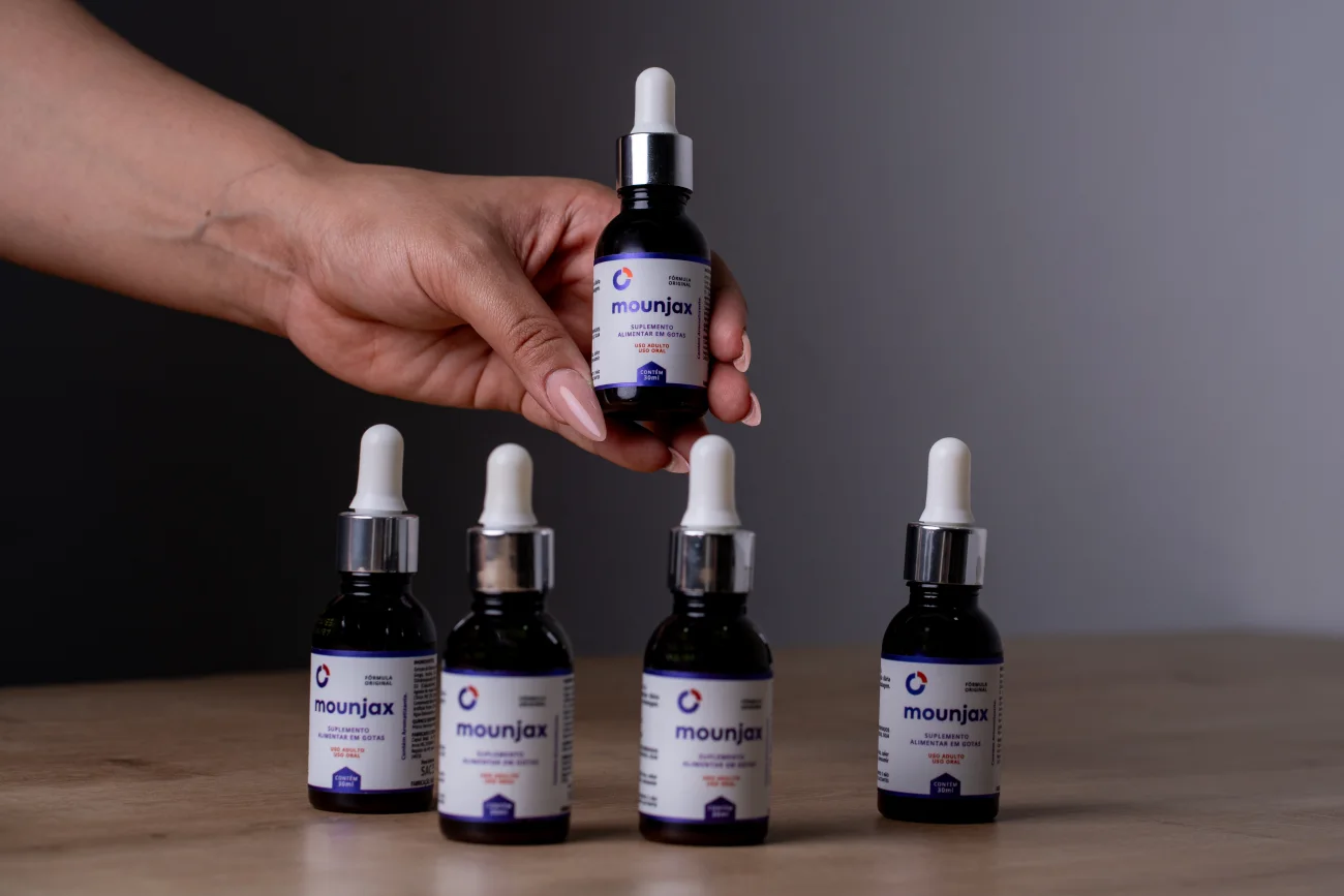Uma m&atilde;o pega um frasco de Mounjax de 30 ml de uma fileira de quatro, sobre mesa de madeira.